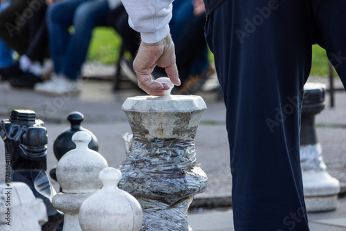 Fotografi Hand gripping a large outdoor chess piece above the board, capturing a decisive moment in gameplay with shallow depth of field and blurred background spectators