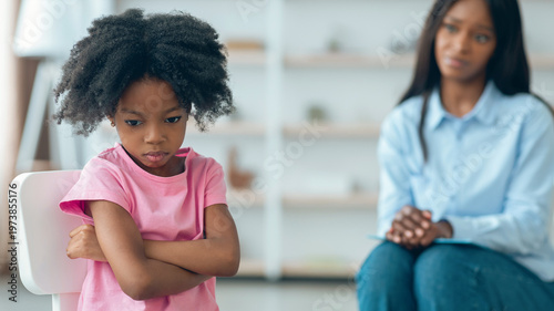 Εκτύπωση καμβά A child with curly hair sits on a chair with crossed arms, showing signs of frustration while an adult sits nearby