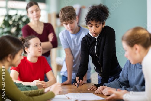 At round table conference, teen participants solve problems, try to create task solution plan, teacher watching. Brain storm, psychological training, interaction between students and teachers