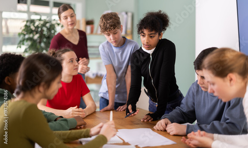 Group of teenagers and students discuss the task at the table, they exchange opinions and theories against the background of teacher in classroom. Schoolchildren look at the paper and solve the task