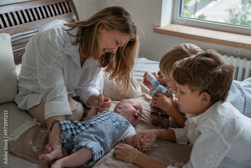 Картината върху платно Young mother and two children play with a newborn baby on a bed in a bright home