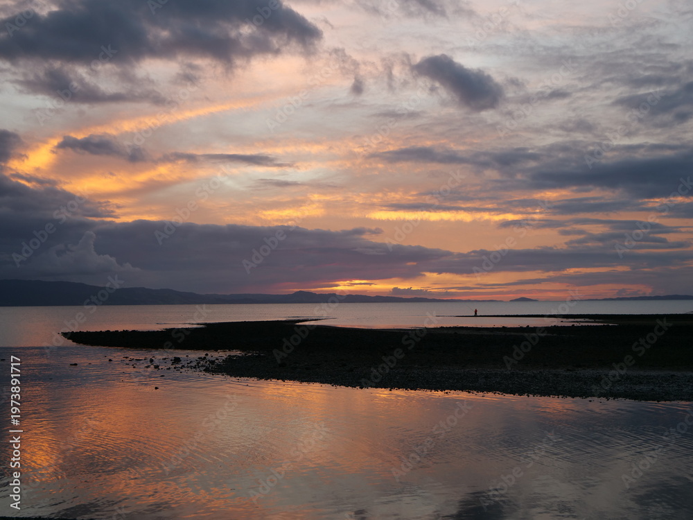 Obraz premium Serene coastal sunset over calm waters and silhouetted hills. Dramatic orange and purple clouds reflecting on the tide at dusk. Peaceful evening landscape in New Zealand with a lone fisherman.