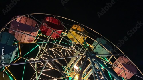 Close-up shot of a colorful Ferris wheel at night with vibrant green, yellow, and orange lights against a dark background