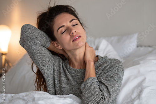 Woman rests in bed, eyes closed, hand on neck. Soft light glows nearby, peaceful atmosphere. Comfortable sweater suggests relaxation or recovery. Ideal for health, wellness, or stress relief content
