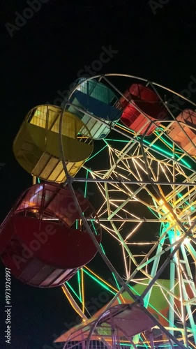 Close-up shot of a colorful Ferris wheel at night with vibrant green, yellow, and orange lights against a dark background