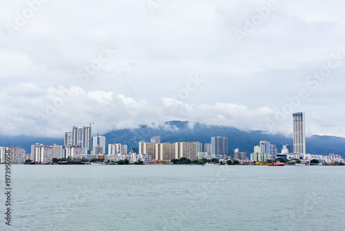 Penang skyline along waterfront. Modern high-rise buildings stretch beside calm sea. Aerial coastal cityscape view under cloudy sky.