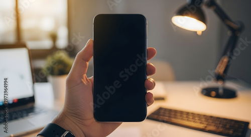 Male hand holding dark smartphone with black screen in office environment with laptop desk lamp and keyboard.