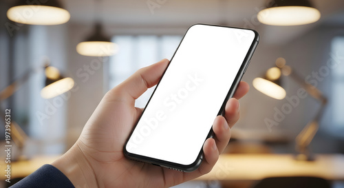 Hand holding modern smartphone with blank white screen in contemporary office workspace with lamps and bokeh lights.