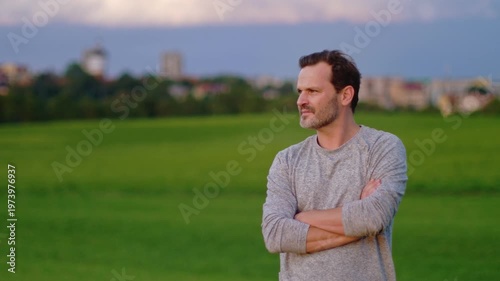 Portrait of a confident man standing out in green field at sunset with rural town in the background