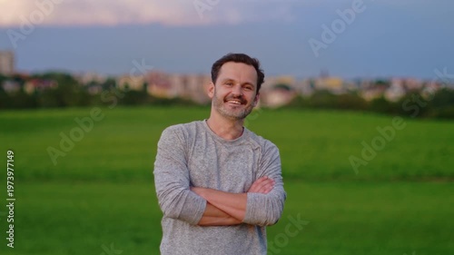 Video Testimonial portrait of a middle aged man in casual dress standing out in a green field with town architecture blurred in the background