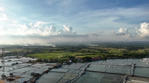 Aerial View of Traditional Salt Evaporation Ponds Bordering Green Rice Fields and Village Settlement under Cloudy Sky