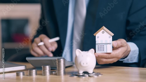 Businessman calculating home loan costs with calculator, coins, and piggy bank, then showing house model.