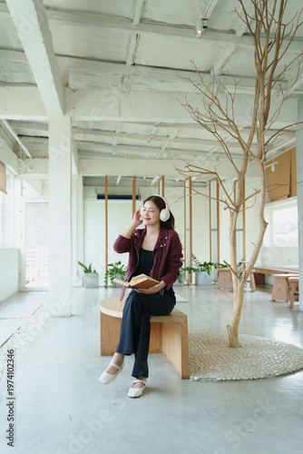 Asian woman relaxing on a bench, listening to music with white headphones and holding a book in a modern and minimalist cafe environment with a decorative indoor tree