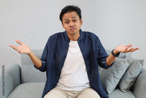 Tablou pe pânză An young man sits on a sofa at home while shrugging his shoulders and opening his palms, with a confused, questioning expression, looking at the camera
