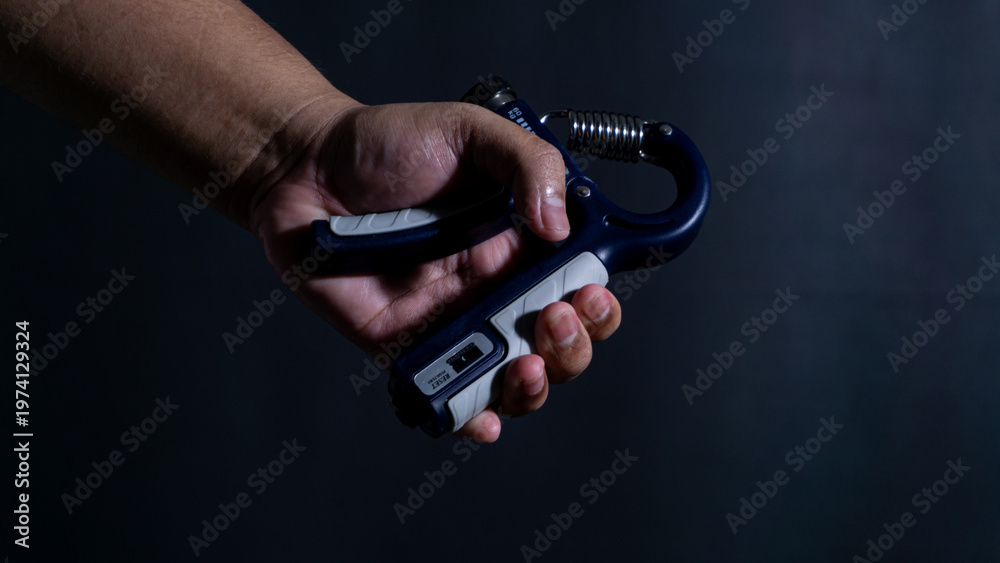 Fototapeta premium A male hand tightly clamps down on a blue and grey hand strengthener in front of a black background, indicating an intense grip training session. The single light source creates heavy
