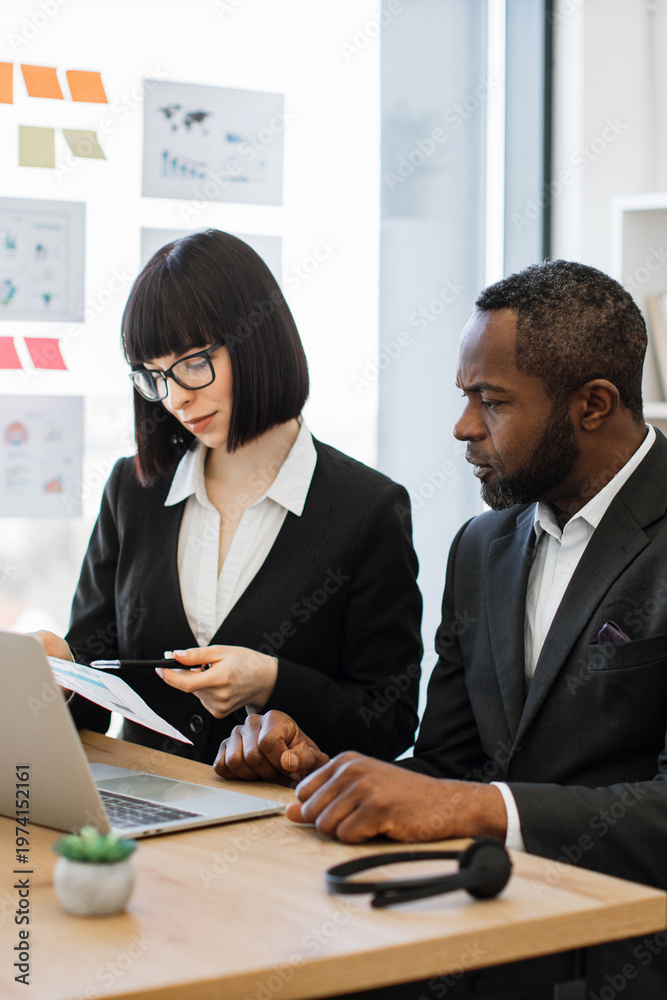 © sofiko14 - A woman in glasses and a man in a suit collaborate on a project at a desk with a laptop and charts on the wall