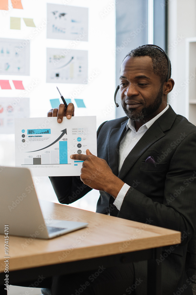 Fototapeta premium An African American man in a suit wearing a headset points to a bar graph showing upward growth during a virtual meeting