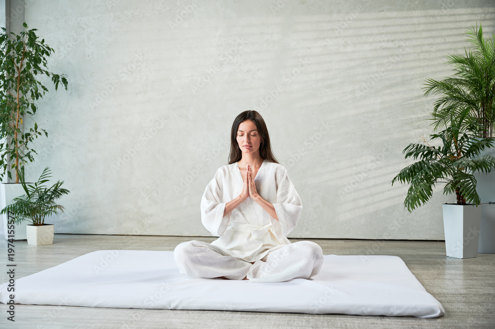 Obraz premium Beautiful woman in white dress sitting on mat in lotus position before Thai massage, practicing yoga, showing namaste sign.
