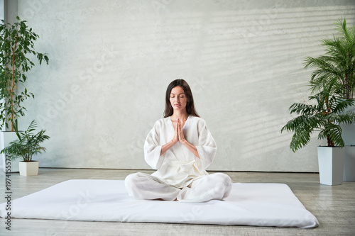 Beautiful woman in white dress sitting on mat in lotus position before Thai massage, practicing yoga, showing namaste sign.