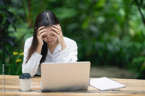 Young professional woman with stress headache sitting at a desk with laptop, coffee cup, and notebook in a serene green outdoor environment