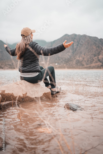 Woman in puffer jacket and cap meditating by a lake with mountains in the fog