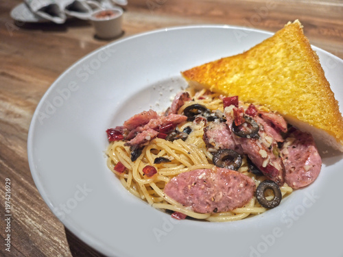 Spaghetti Aglio e Olio with sliced sausages, black olives, and dried chili, served with a piece of crispy golden garlic bread in a white bowl