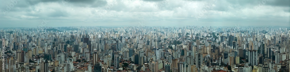 Fototapeta premium Cityscape Panorama under Cloudy Sky: Capturing the vastness and density of an urban landscape, rows of skyscrapers and buildings stretch towards the horizon under a dramatic, cloudy sky.
