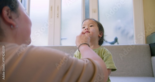 Photos Asian toddler patiently receives gentle facial application of skincare cream in a bright living room, focusing on pediatric dermatology, daily hygiene routine, and tender family bonding