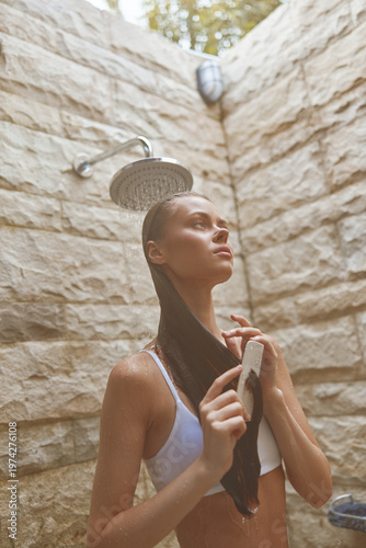 young woman washing long hair under outdoor shower with stone wall background, natural light, summer relaxation, healthy hair care.