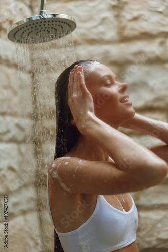 Young woman enjoying an outdoor shower with water flowing over her face and shoulders against a natural stone wall background in warm daylight.