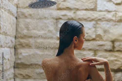 woman showering under outdoor rainfall showerhead with stone wall background, wet hair, relaxed mood, natural lighting, serene wellness moment
