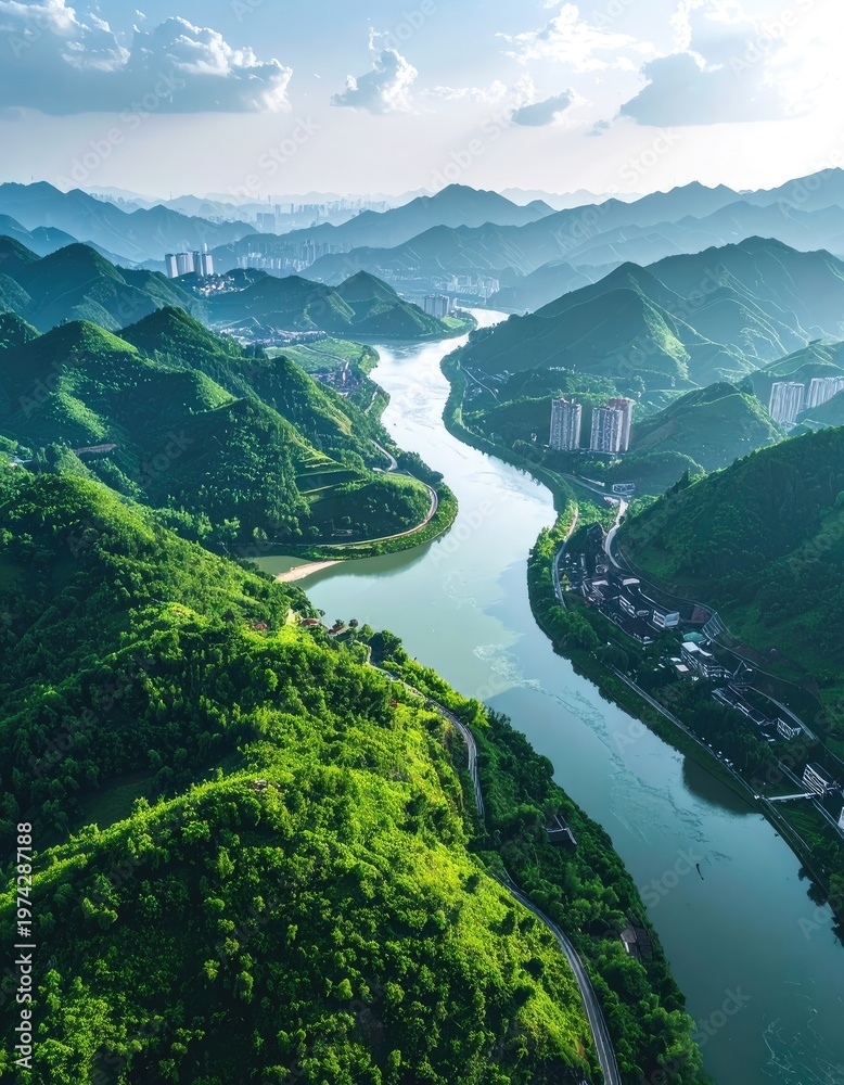 Obraz premium An aerial view showcases a winding river gracefully carving its path through lush, green mountains, with a distant cityscape subtly blending into the horizon under a bright, partly cloudy sky.