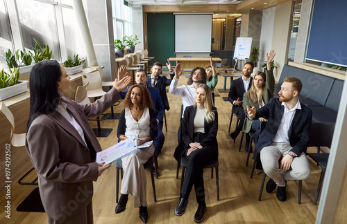 Speaker leads business training for corporate team audience. Employees raise hands during seminar in an office meeting, guided by a manager and coach. Clear concept of teamwork and growth.
