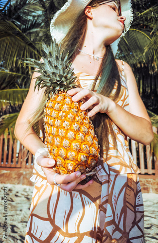 Young woman holding ripe pineapple on tropical beach among palm trees. Concept of summer vacation, exotic fruits and relaxed island lifestyle