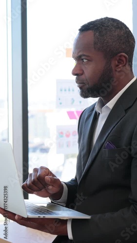 An African American businessman in a suit looks intently at his laptop screen while standing near a window with a city view