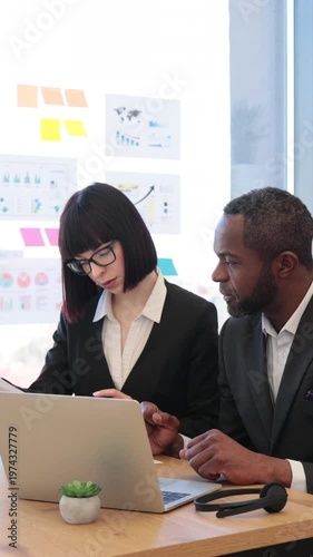 A woman in glasses points at a laptop screen while discussing data with an African American man in a suit during a business meeting