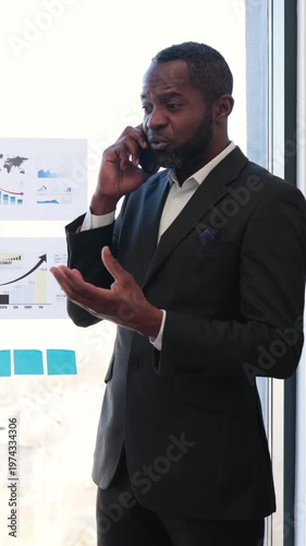 A concerned executive gestures while discussing charts and financial reports during a mobile call in a bright, glass-walled office