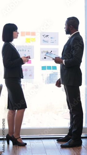 Two professionals, an African American man and an Asian woman, collaborate on a project using charts and sticky notes on a glass wall