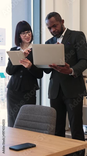 Two professionals consult a laptop and tablet in a modern office by a window while preparing a presentation for a meeting with colleagues and stakeholders