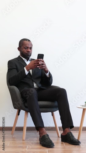 An African American man dressed in a formal suit sits in a modern chair while intently focused on his mobile phone