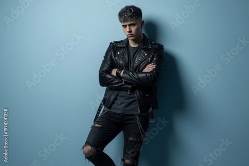 Young person stands against blue wall wearing black leather jacket and ripped jeans while posing confidently and showing a serious expression