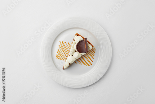 Top view of a gourmet tart slice with whipped cream, chocolate garnish, and caramel drizzle on a white plate. Minimalist aesthetic on a white background.