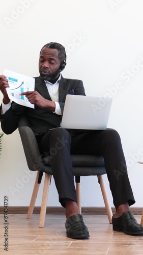 An African American man in a suit sits in a chair, holding a document with a graph and pointing to it while wearing a headset and looking at a laptop