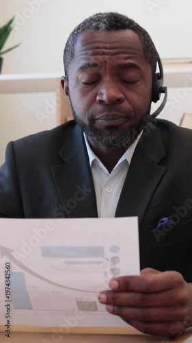 An African American man wearing a headset and suit examines a document with a rising graph, indicating business growth and analysis