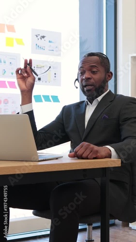 An African American man wearing a headset and suit speaks animatedly while on a video conference call with a laptop in front of him