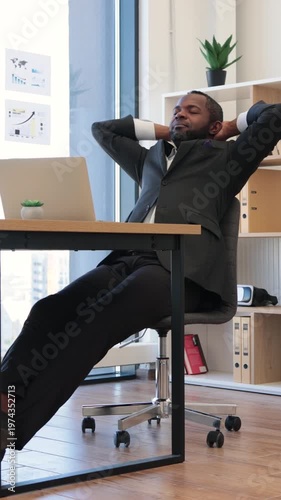 An African American man in a suit leans back in his office chair with his hands behind his head, looking content and successful