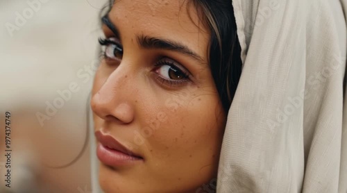 Young Middle Eastern woman with dark hair and brown eyes, wearing a light-colored headscarf. Natural background with soft focus.