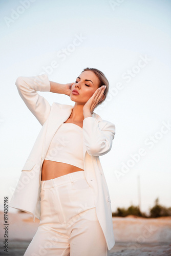Slika na platnu Woman in white suit with hands on temples, serene expression against a soft sky