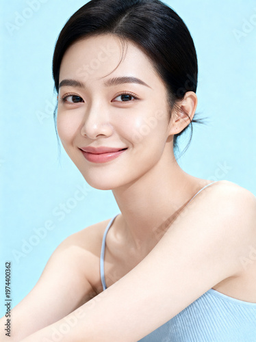 Close-up portrait of a beautiful young Asian woman with glowing skin smiling gently against a soft blue background.