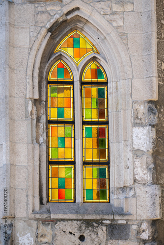 Colorful stained glass window set in a Gothic stone frame, highlighting geometric patterns, weathered masonry and historic architectural detail.